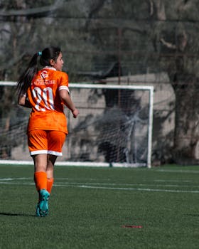 A female soccer player in orange uniform runs towards the goal on a lush green field.