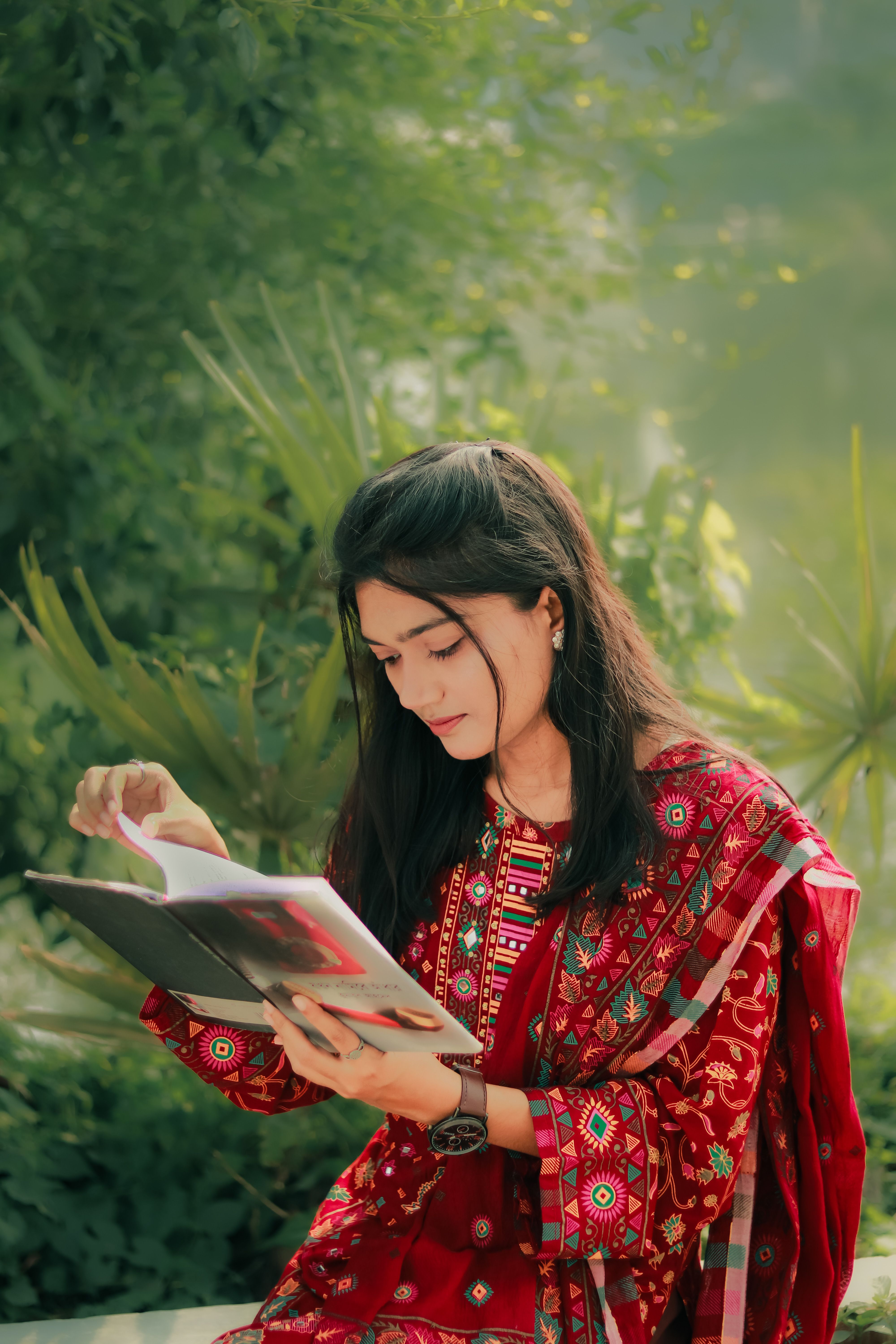 A woman in traditional clothing reads a book outdoors in a lush garden setting in Bangladesh.