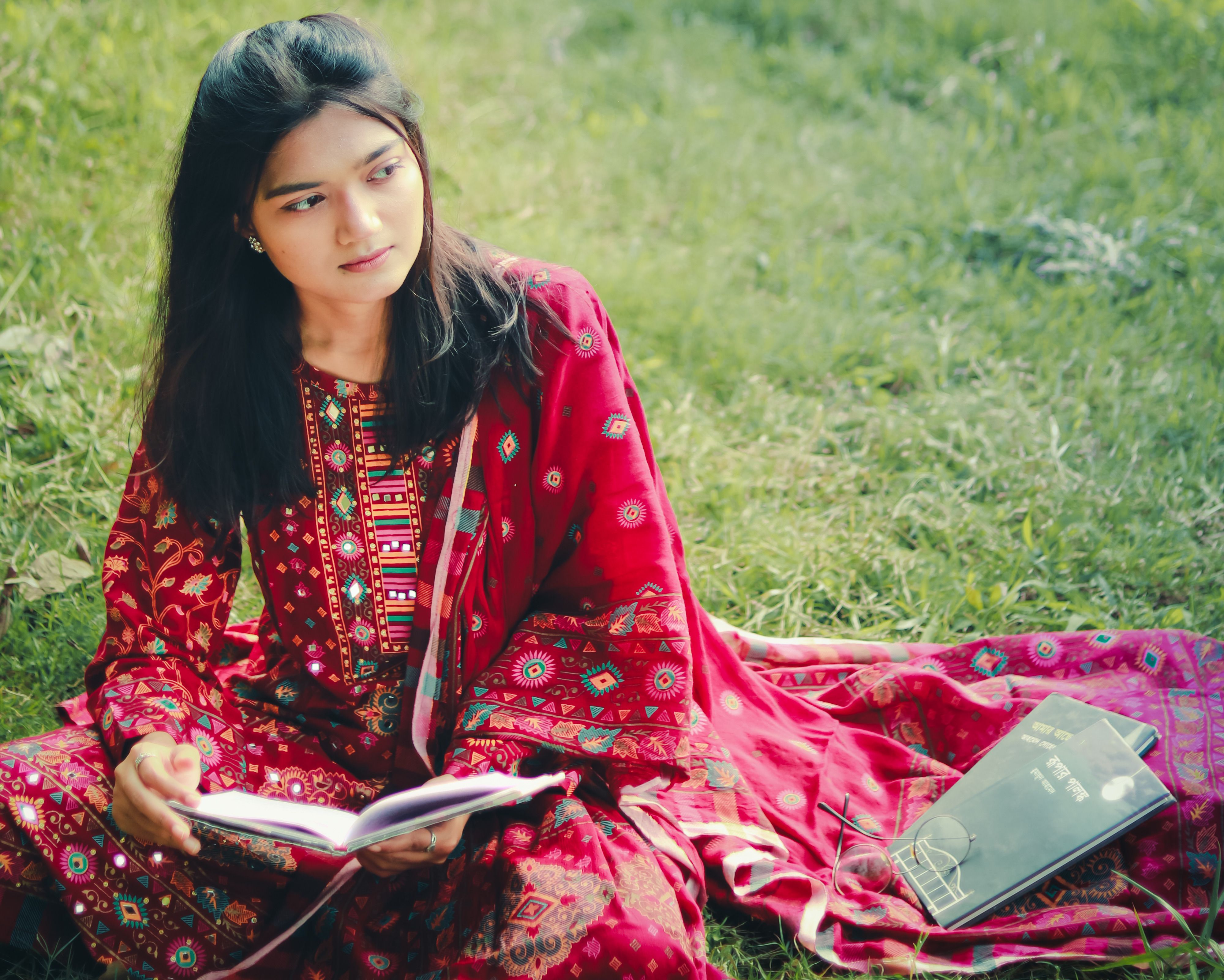 A young woman in a vibrant traditional outfit reading a book while sitting on grass in Khulna, Bangladesh.