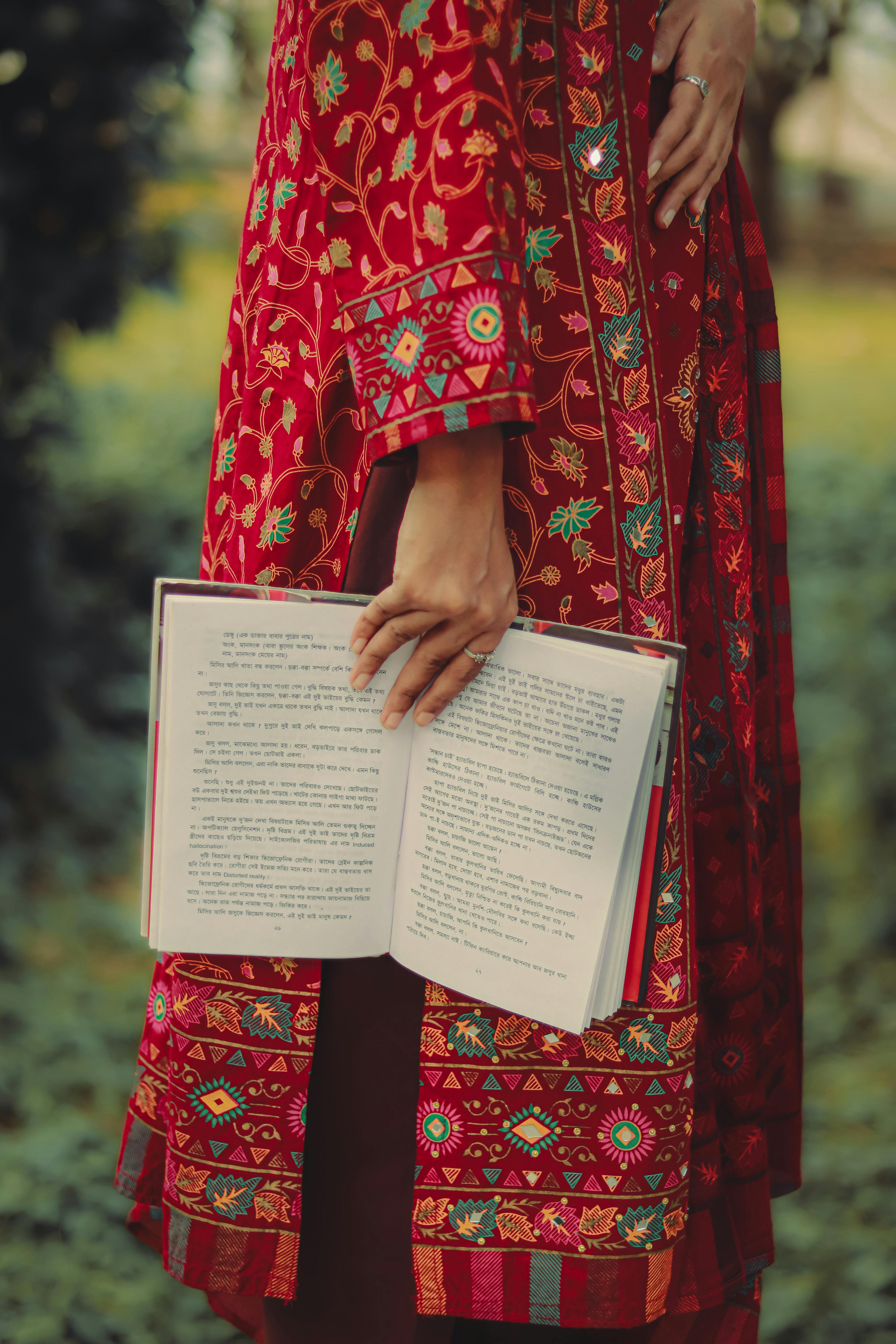 A woman in vibrant ethnic attire holds an open book outdoors, illustrating contemplation and culture.