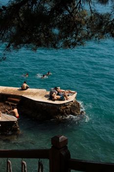 People sunbathing on a scenic deck by a clear blue sea under a tree.