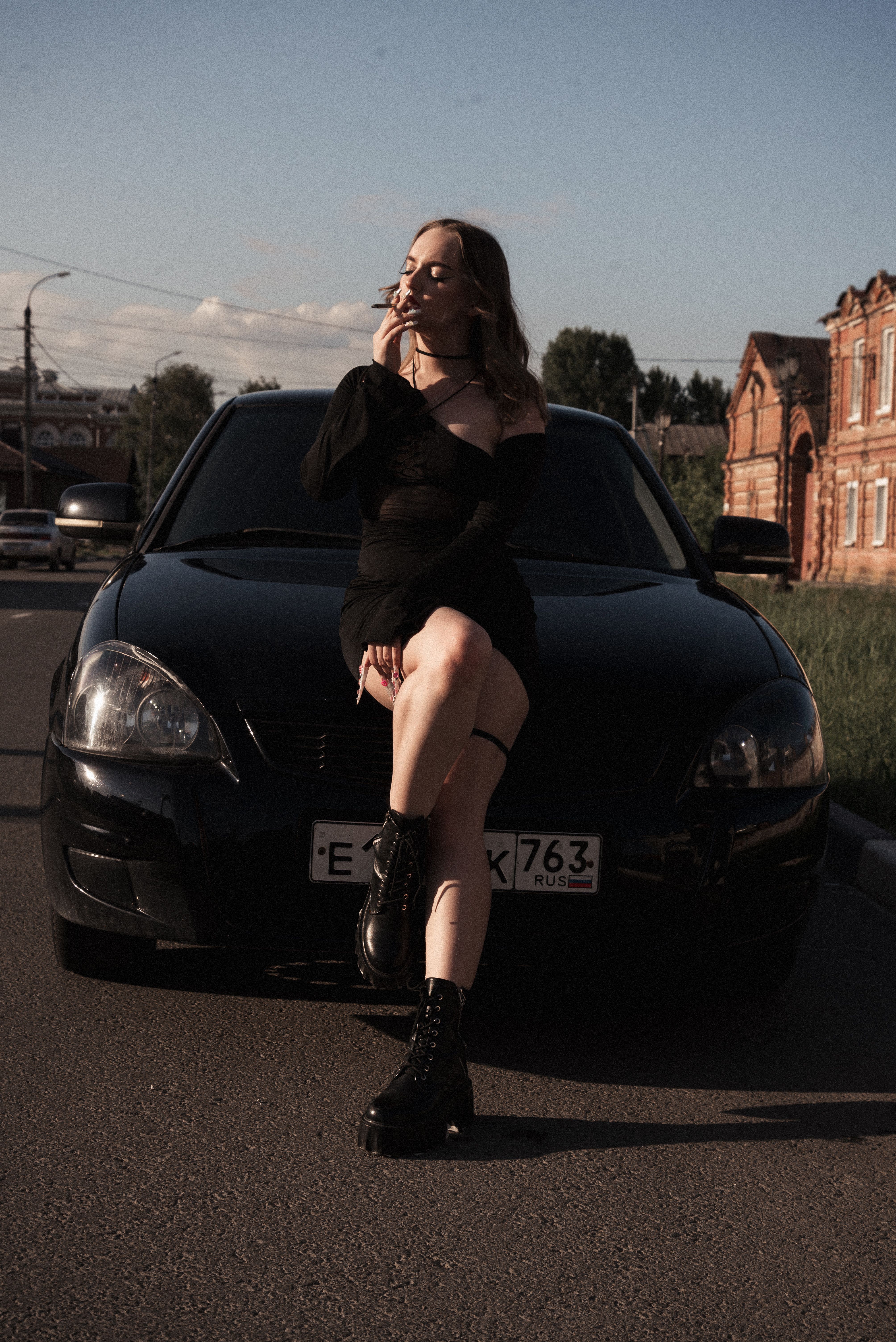 Stylish woman in black dress seated on a car hood, street setting at sunset.