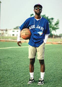 A stylish young man in sports attire holding a basketball on an outdoor field.
