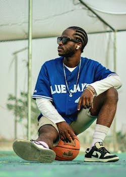 Stylish man in Blue Jays jersey sitting on basketball outdoors. Trendy sports fashion.
