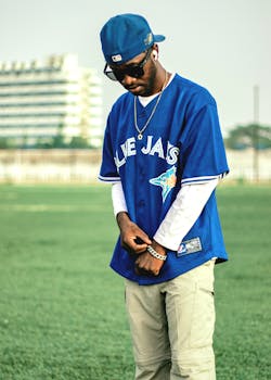 A trendy man in a blue baseball jersey stands confidently on a green field outdoors.