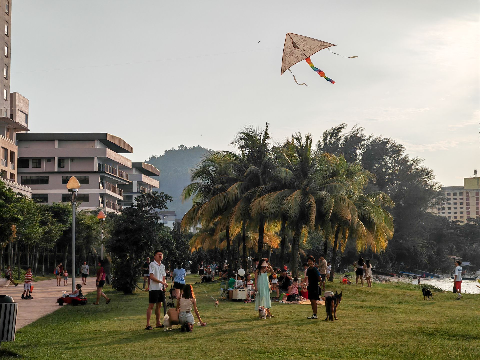 Families enjoying kite flying at a park in Tanjung Bungah, Penang, under a warm sun.