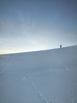 A lone person stands on a snowy slope during sunset, capturing tranquil winter isolation.