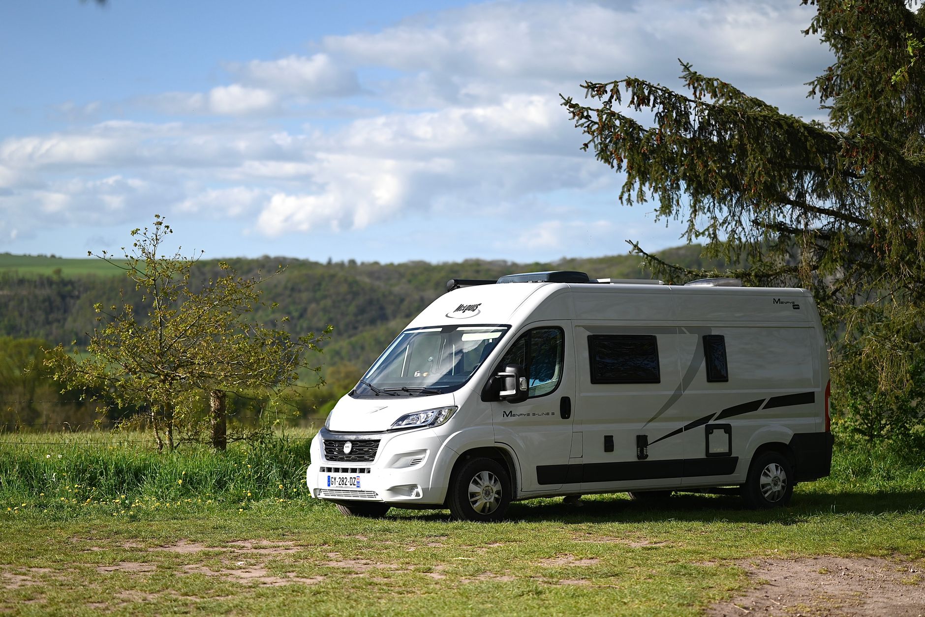 Een witte camper staat op een grasveld, omringd door bomen en onder een blauwe lucht.