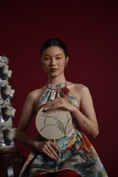 Stylish woman in floral dress holding a fan surrounded by stacked teacups on a deep red background.