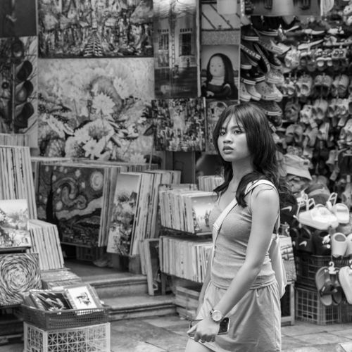 Free A young woman walks by a vibrant street art market, showcasing various paintings and shoes. Stock Photo