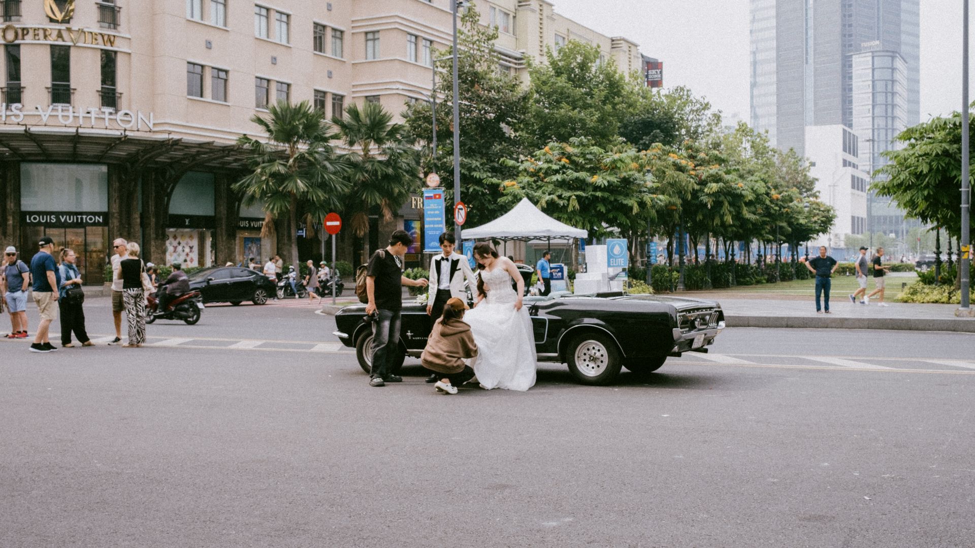 Bride and groom pose by a classic car near a city luxury store, capturing urban wedding vibes.