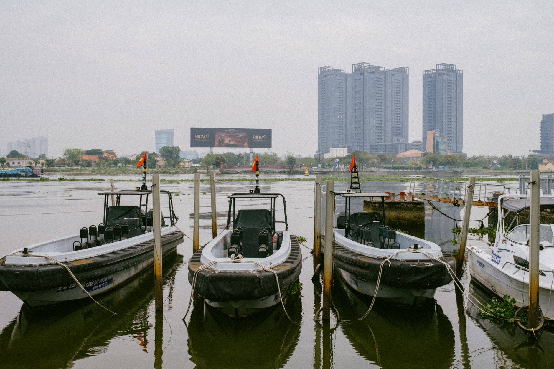 Boats moored along the Saigon River with city skyscrapers in the background on a cloudy day.