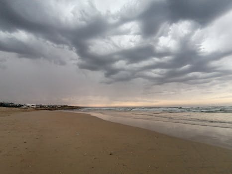 A serene sandy beach under dramatic cloudy skies at dusk, reflecting tranquil ocean waves.
