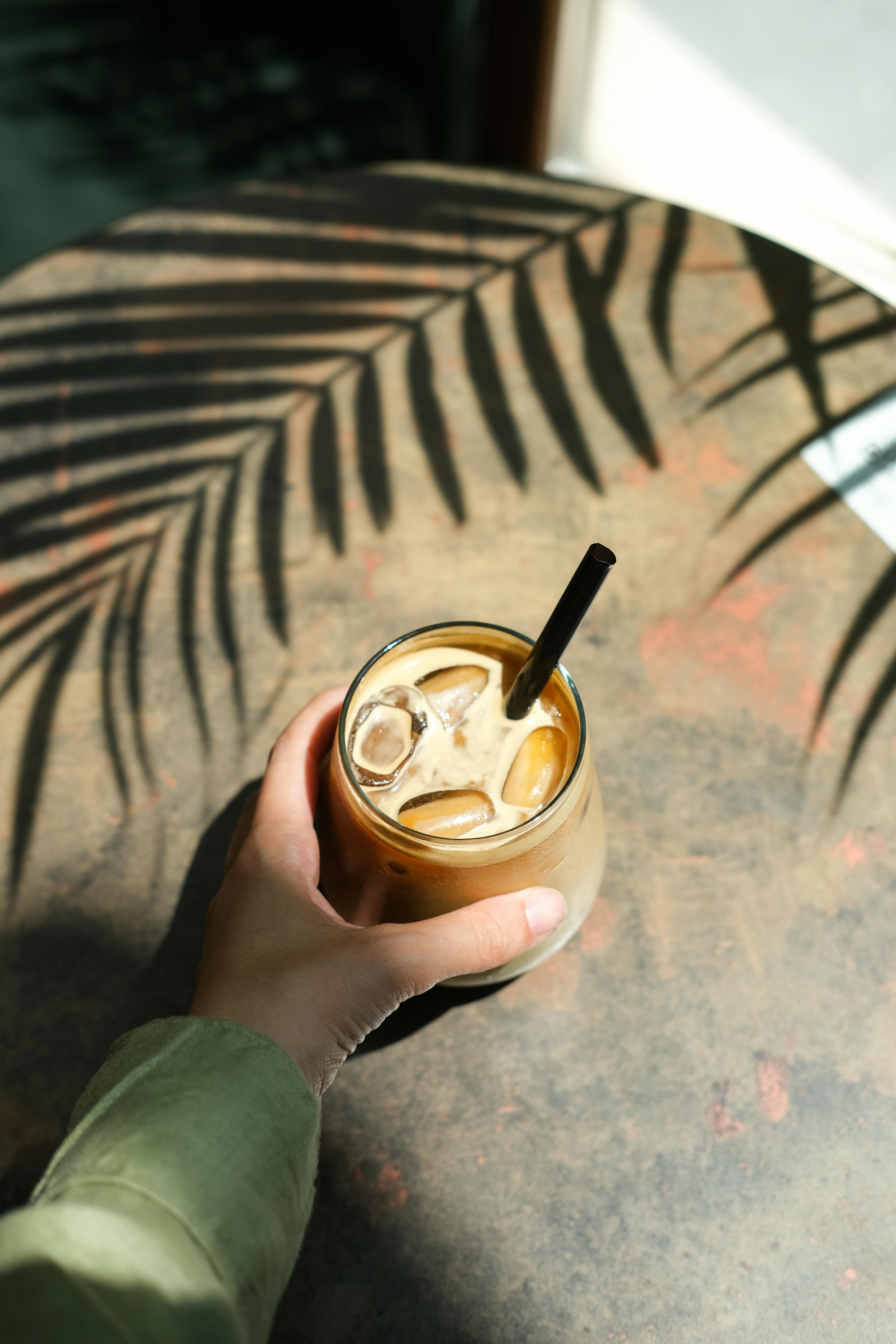 Hand holding iced coffee in glass, on table with tropical leaf shadows in Istanbul cafe.