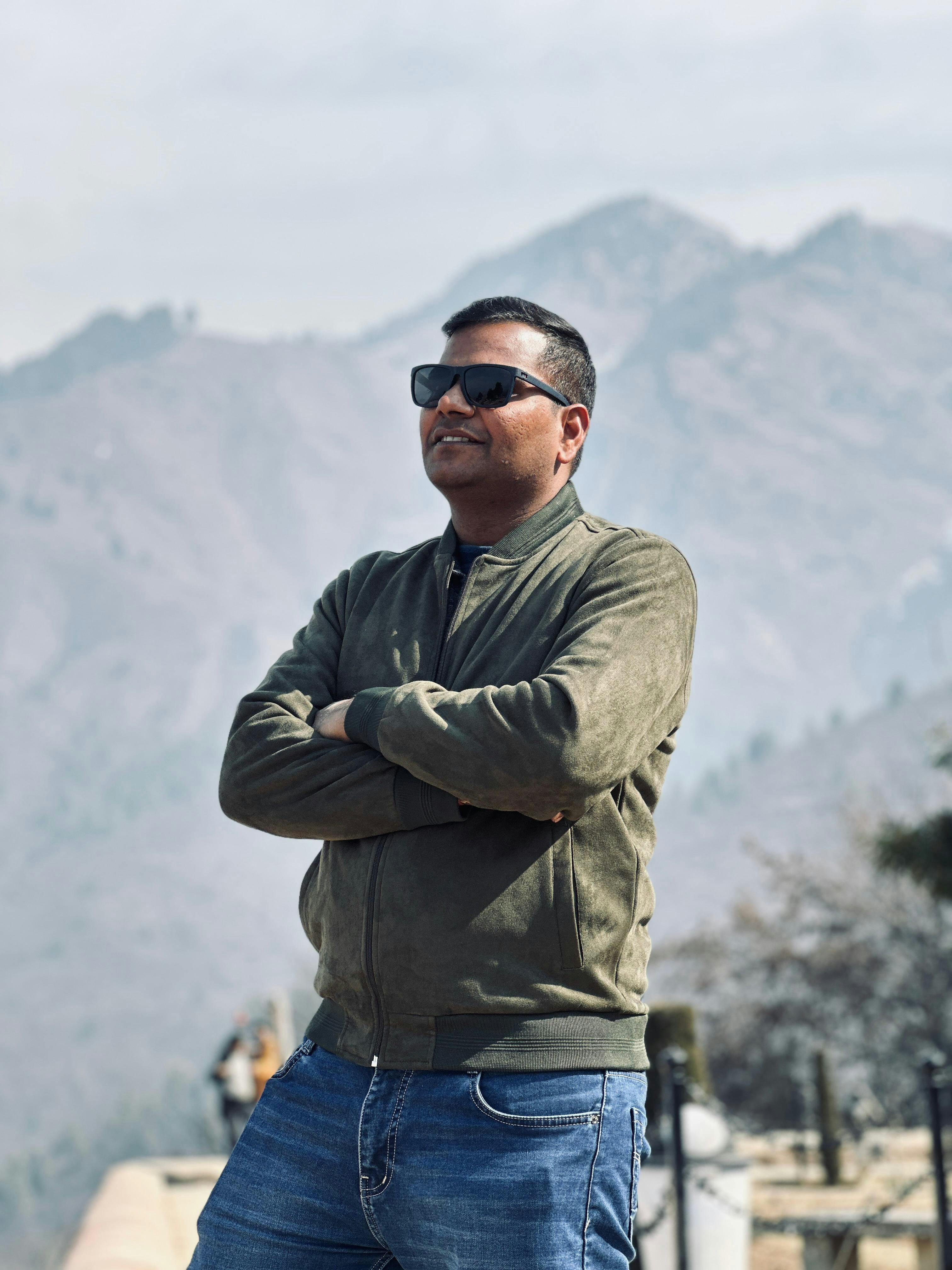 Man in sunglasses and jacket confidently posing with scenic mountain backdrop.