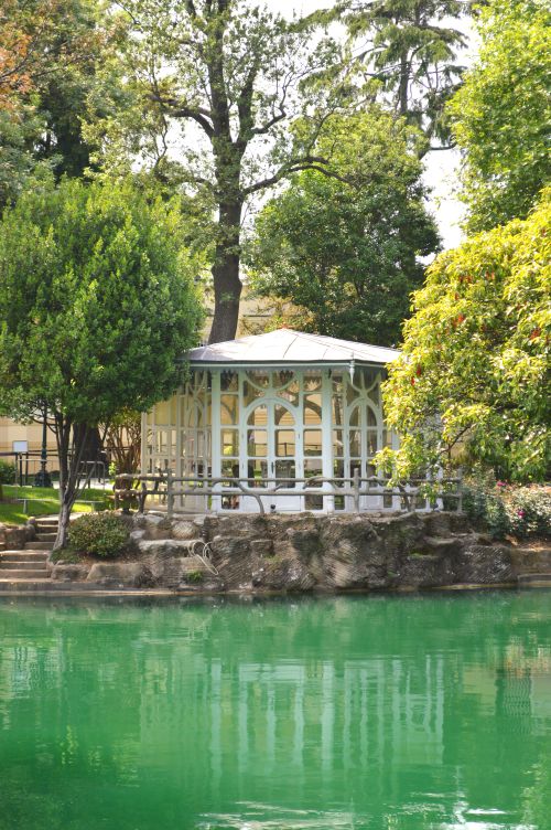 Free A picturesque gazebo surrounded by trees overlooking a tranquil green lake in a park setting. Stock Photo