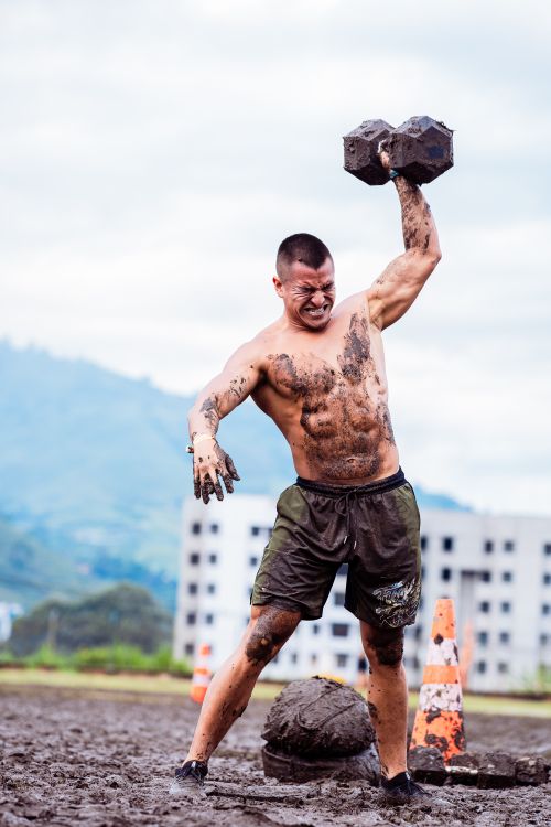 Free A muscular man exercises with a heavy dumbbell in a muddy outdoor setting, showcasing strength and endurance. Stock Photo