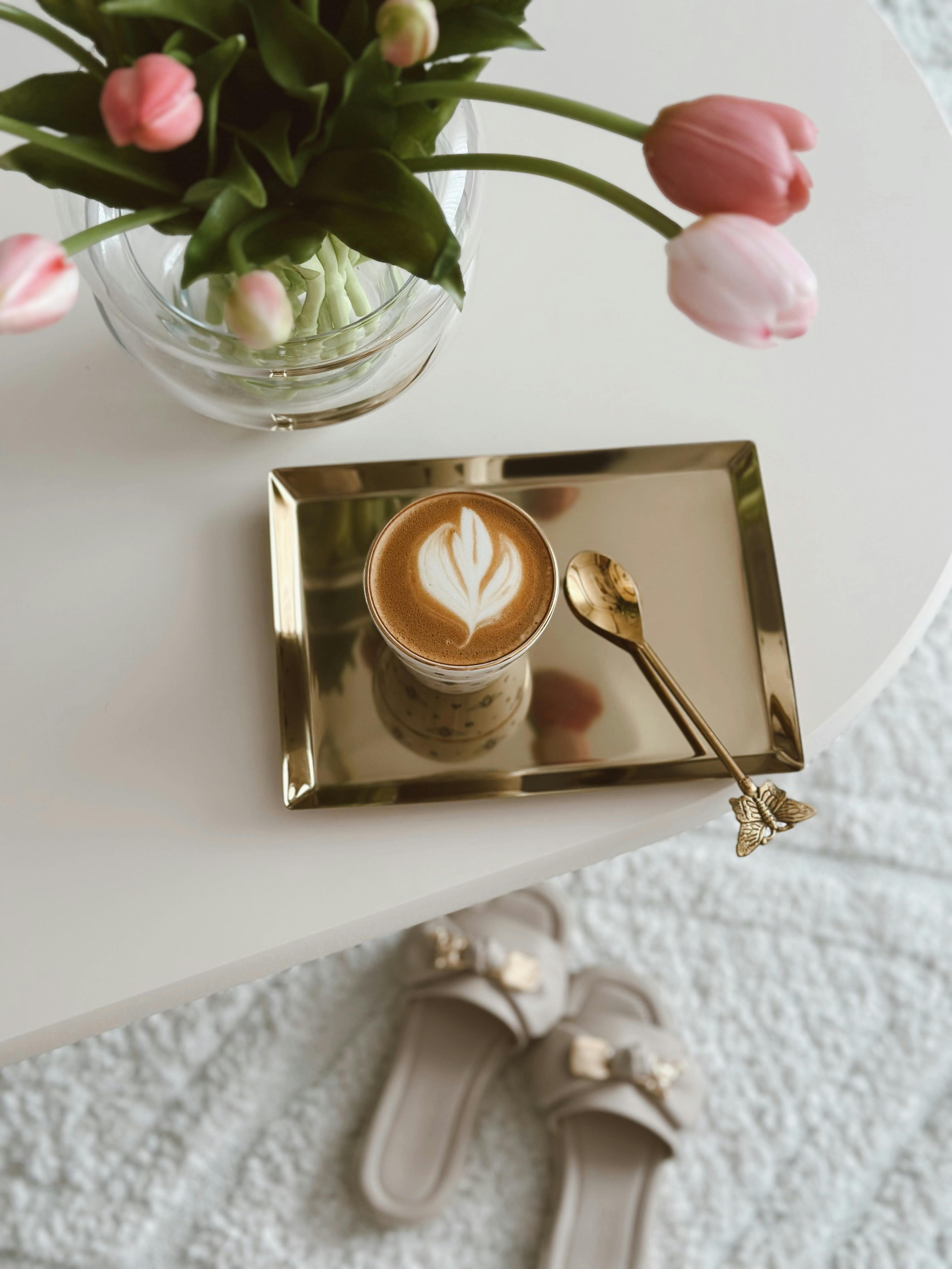 A stylish coffee cup with latte art on a gold tray beside pink tulips. Perfect for lifestyle posts.