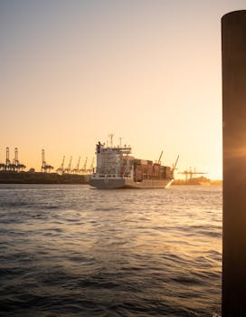 A container ship navigates through Hamburg harbor during a stunning sunset with cranes in the background.