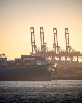 Container ship navigating Hamburg Harbor with cranes at sunset, showcasing global trade.