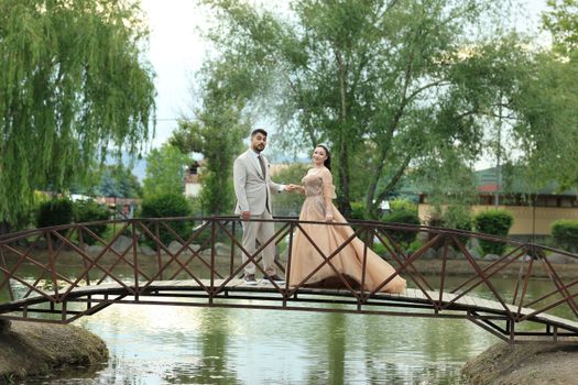 A couple stands on a bridge in a scenic park, dressed in formal attire.