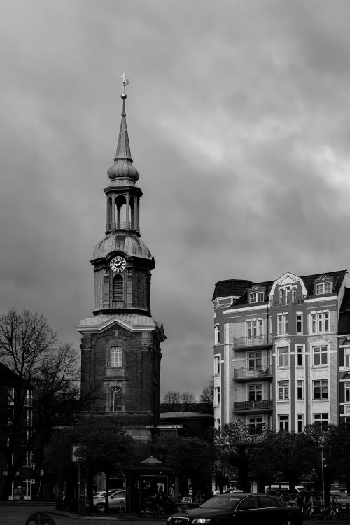 Free Black and white photo of a historic European church tower beside urban buildings under cloudy skies. Stock Photo