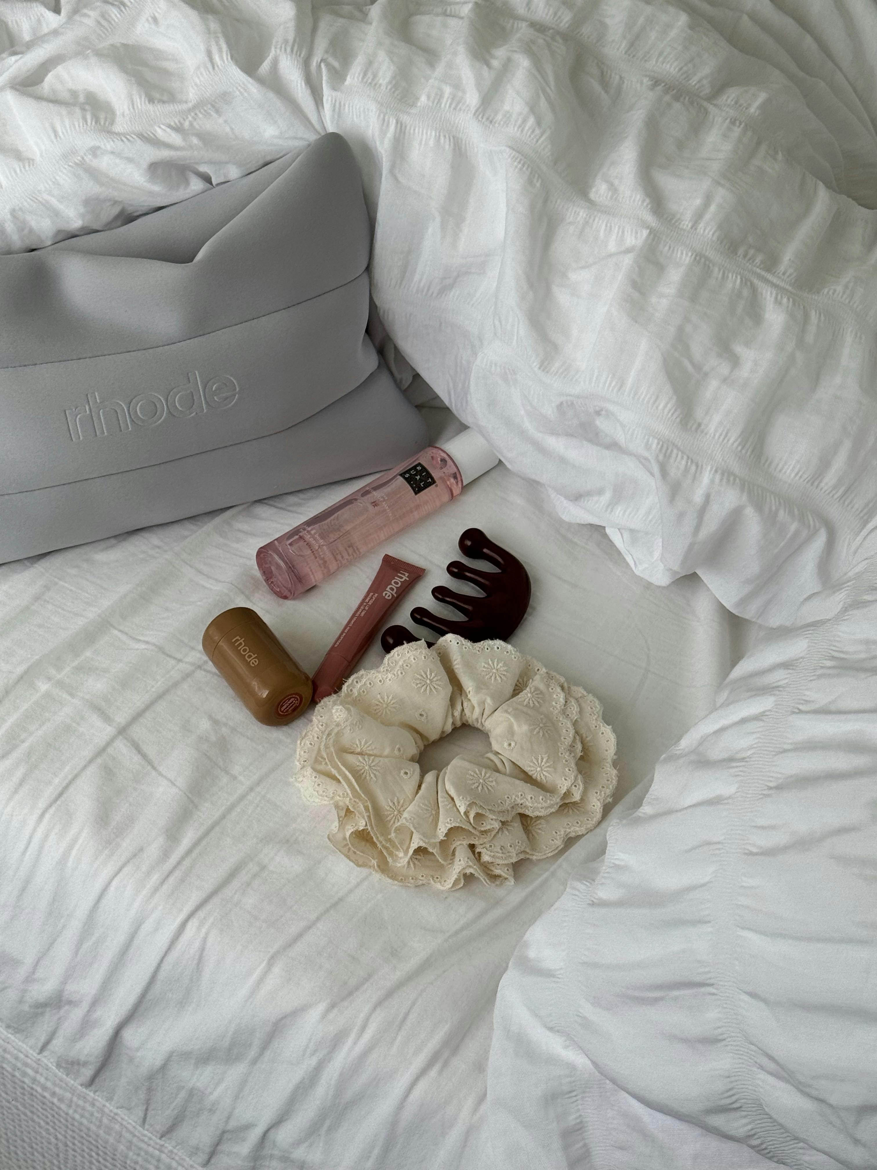Flat lay of beauty products and accessories on a cozy white bed, featuring a makeup bag.
