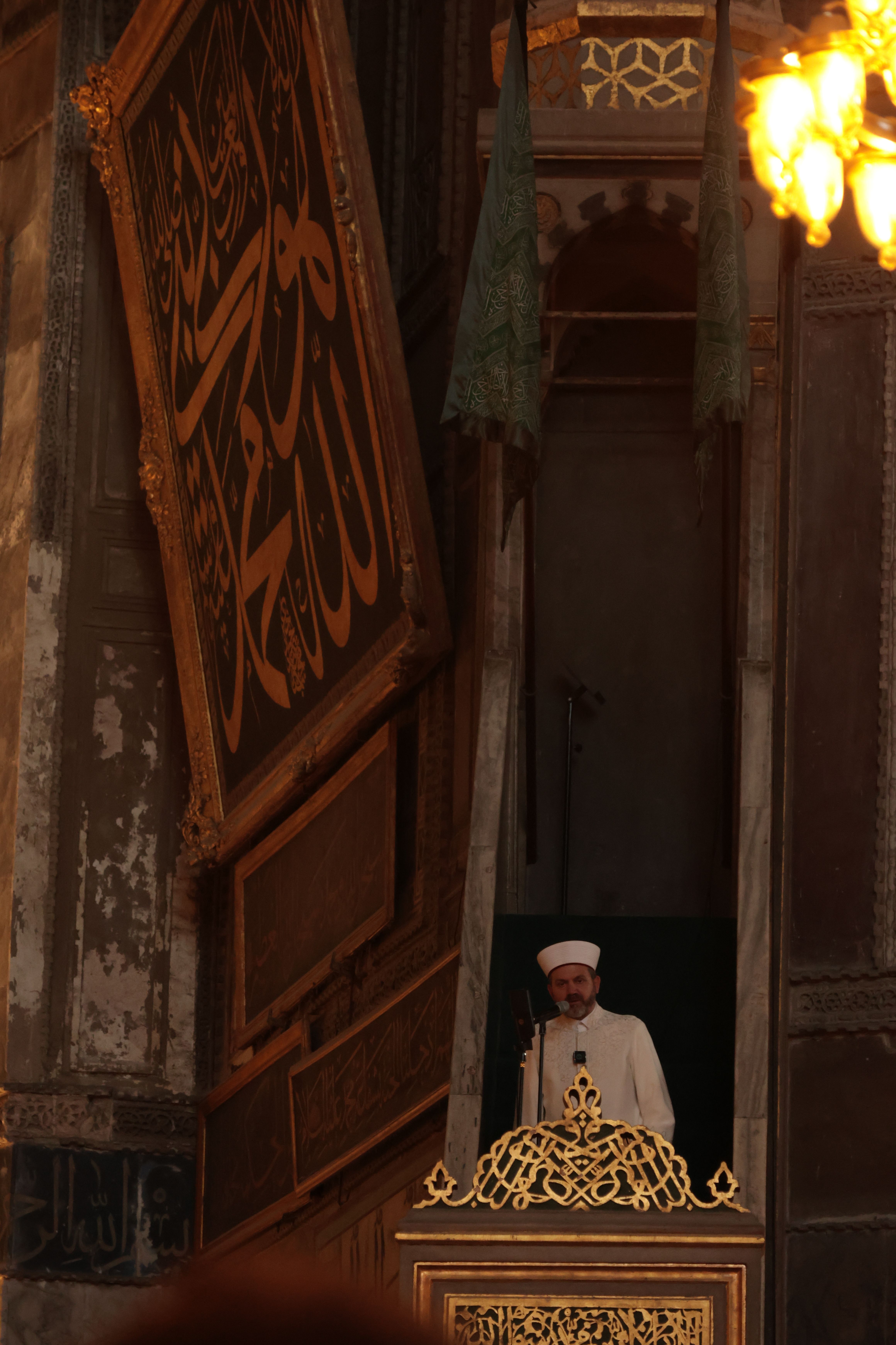 Imam gives a sermon from a pulpit in a historic mosque, ornate calligraphy decorates the walls.
