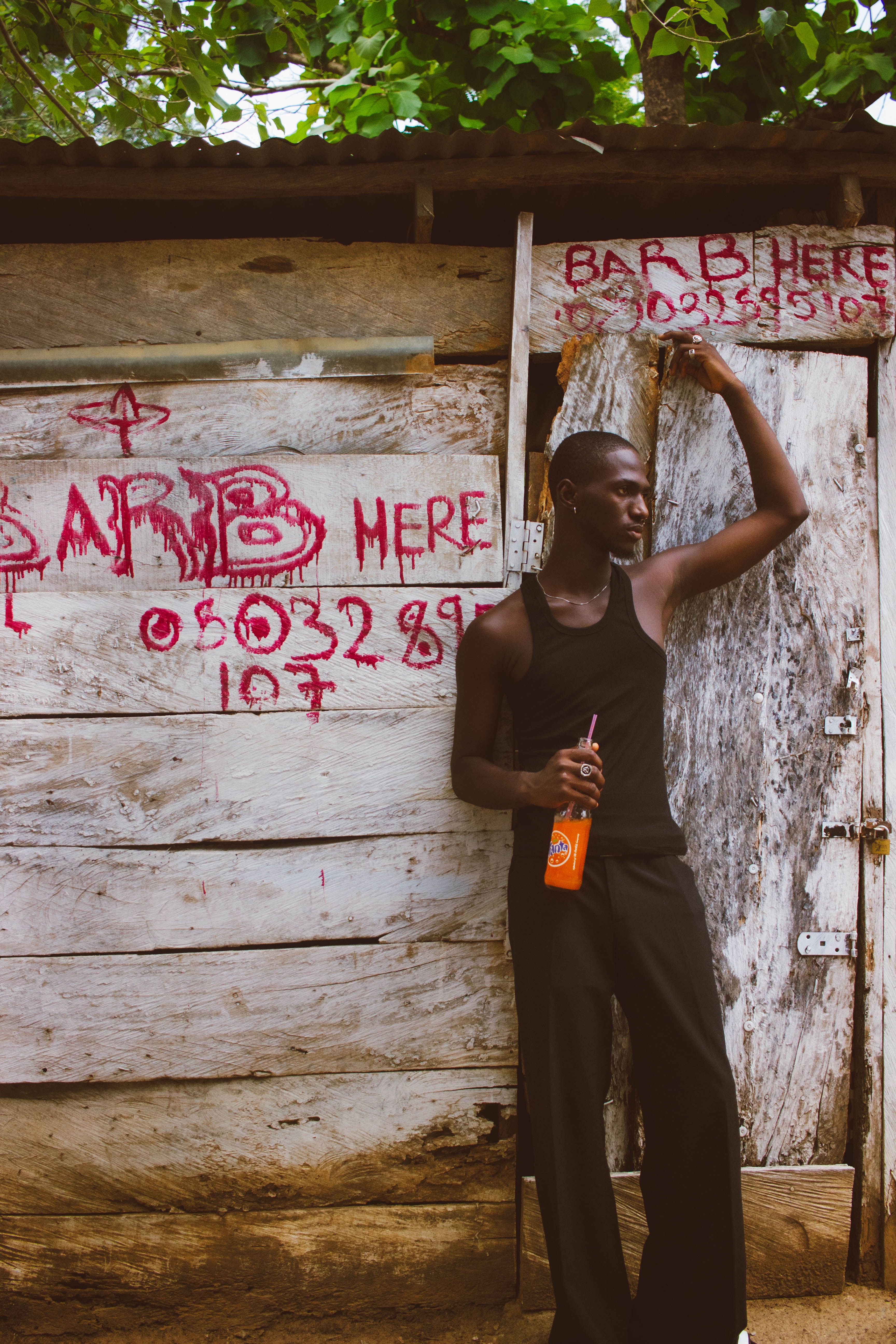 Young man stands casually by a rustic barber shop in Nigeria, holding a drink.