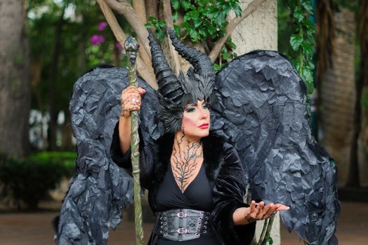 Mysterious woman in a dark sorceress costume with horns and wings, holding a staff in a forest setting.