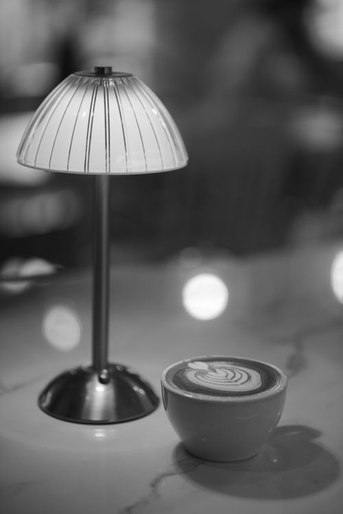 Free Black and white photo of a stylish café scene featuring a cup of coffee with latte art and a table lamp. Stock Photo