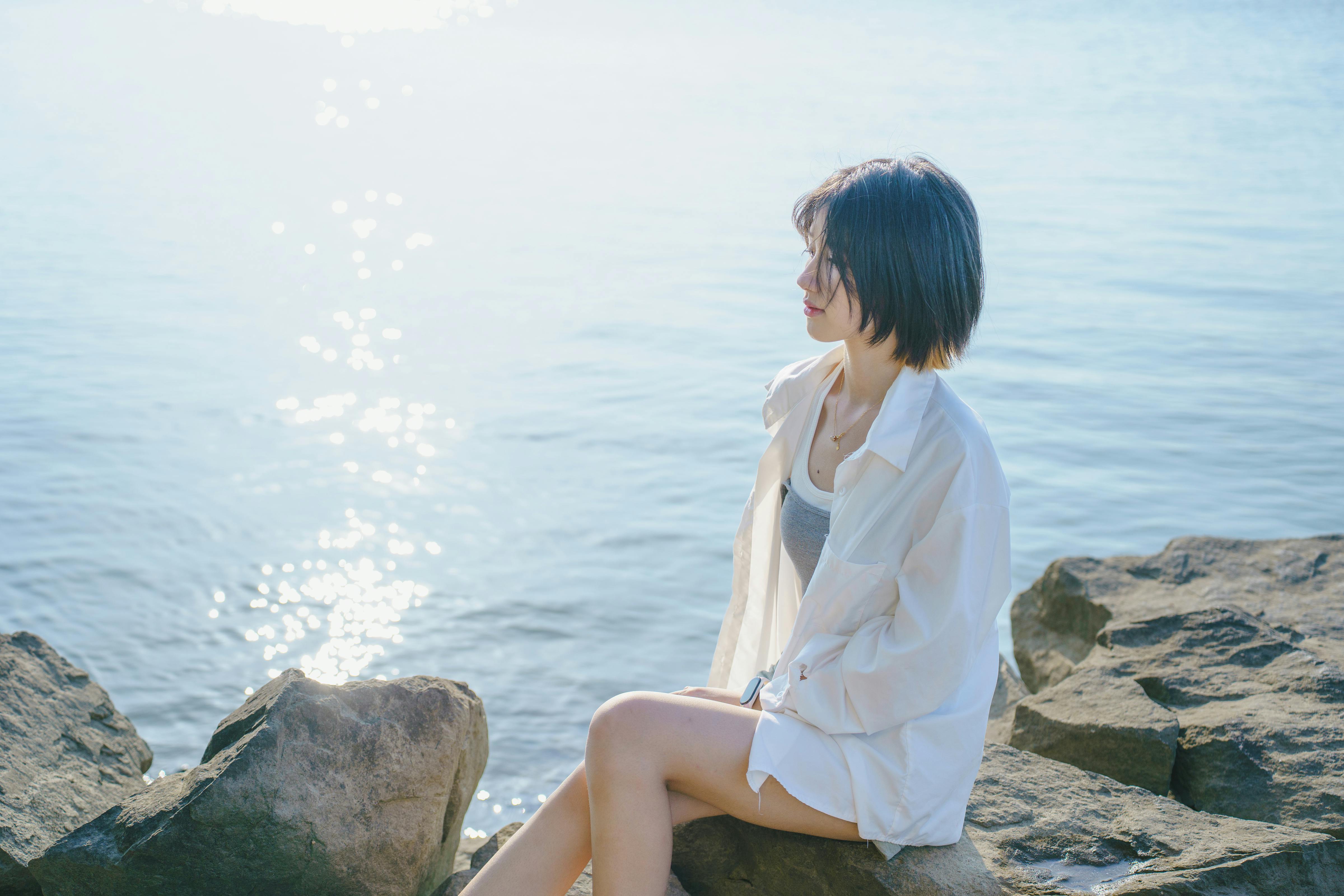 A young woman sits on rocks by the water, enjoying a serene sunrise moment.