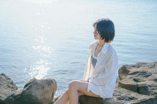Free A young woman sits on rocks by the water, enjoying a serene sunrise moment. Stock Photo