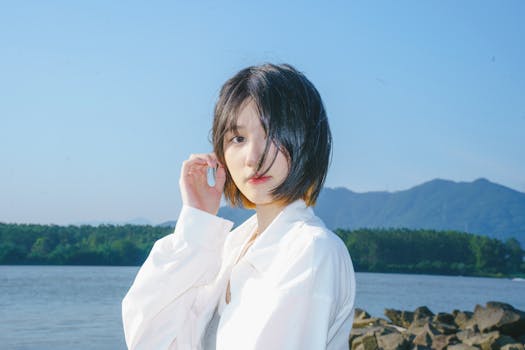Young woman in white blouse posing by river with mountain backdrop on a sunny day.
