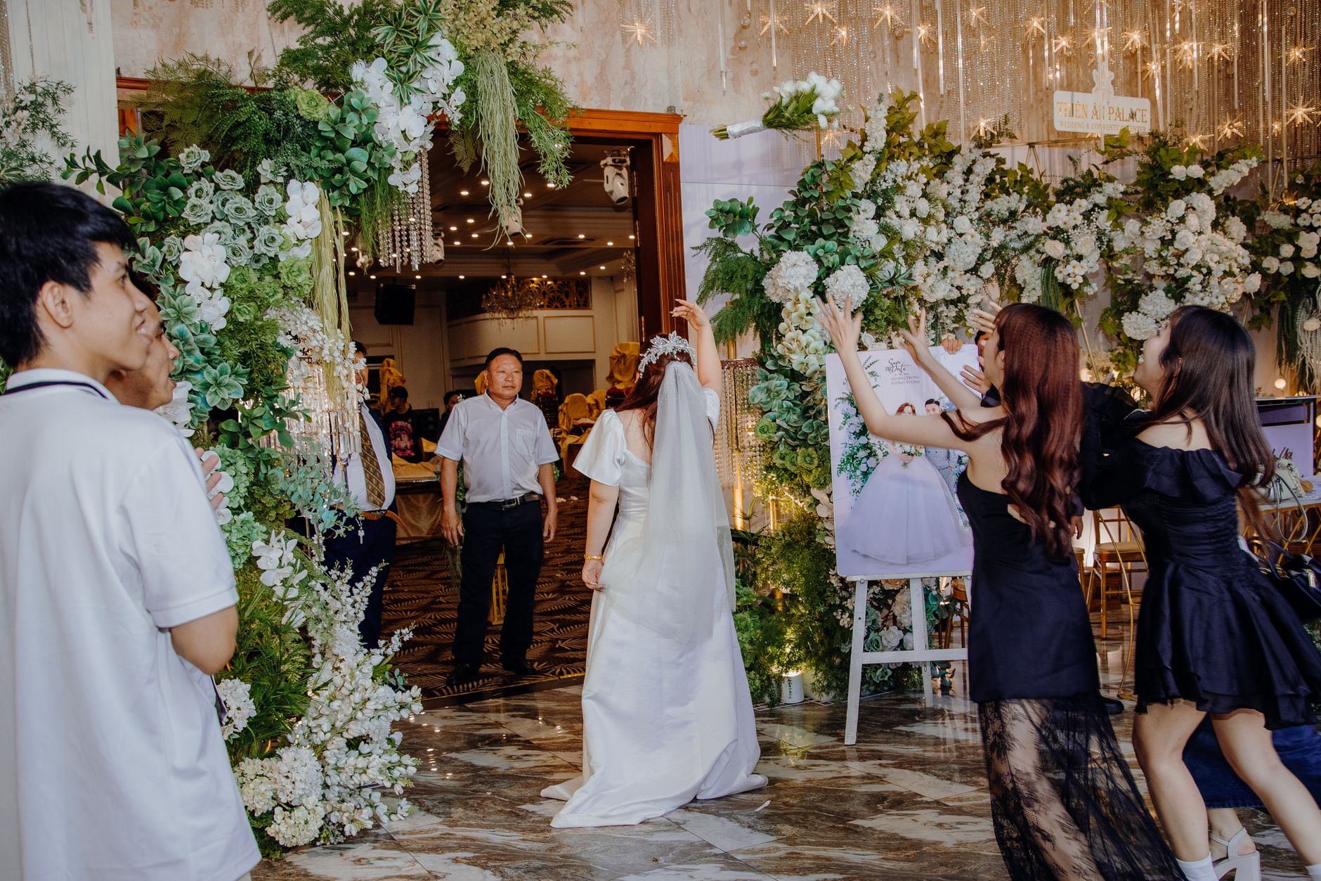 Bride entering decorated wedding venue with joyful guests and floral arrangements.