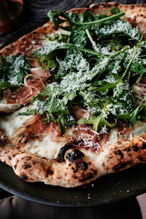 Free Delicious wood-fired pizza topped with fresh arugula and savory prosciutto, perfect for food enthusiasts. Stock Photo