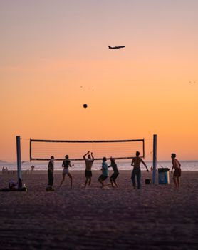 Üzerinde bir uçağın uçtuğu canlı bir gün batımının altında plaj voleybolu oynayan insanların silüeti.