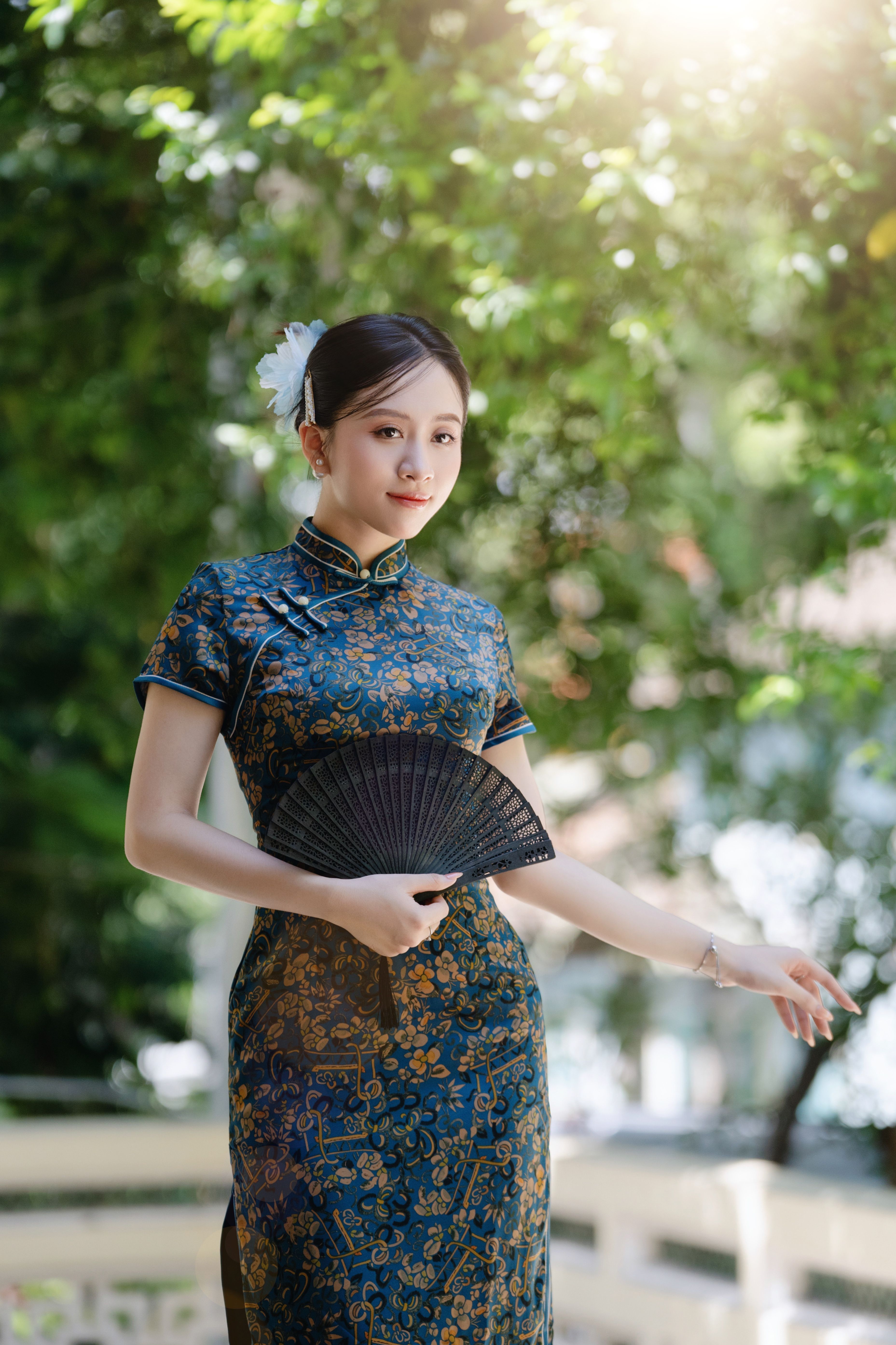 Young woman in traditional Chinese dress with fan, standing outdoors in sunlit garden.