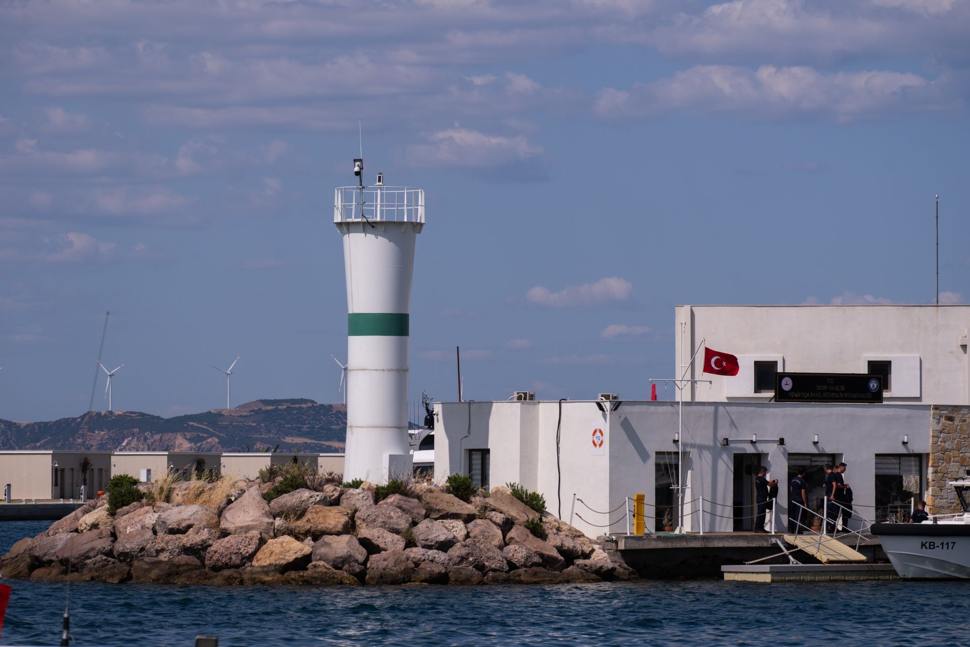 Foça harbor scene featuring a lighthouse, Turkish flag, and distant wind turbines reflecting serene coastal charm.