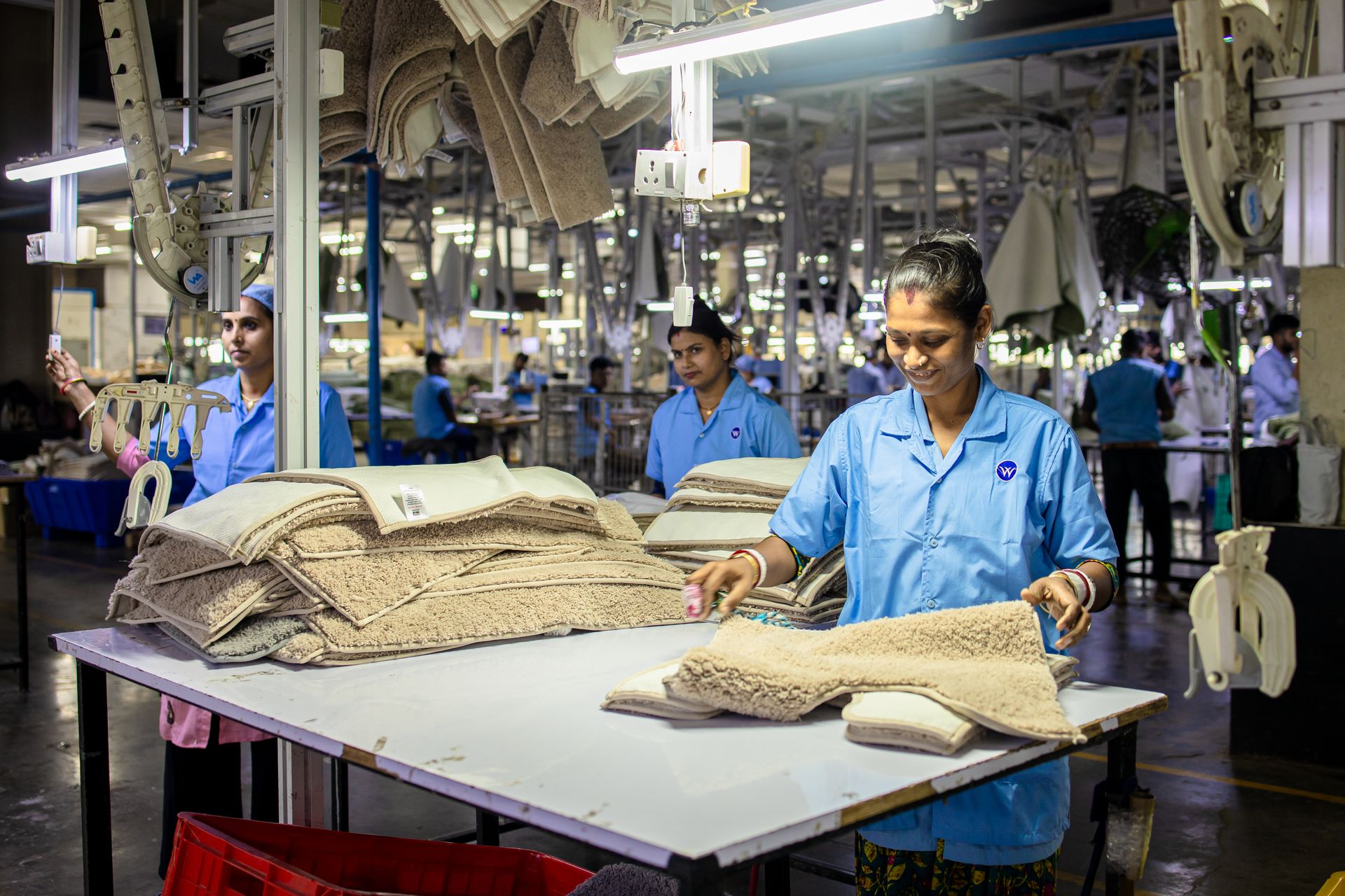 Smiling textile factory workers sorting beige carpet mats in a well-lit industrial setting.