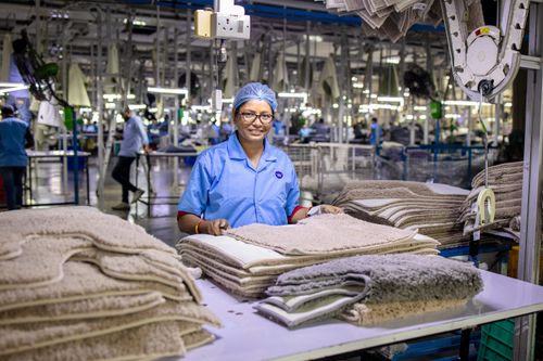 Free A smiling female factory worker stands by stacks of textiles in a modern production facility. Stock Photo