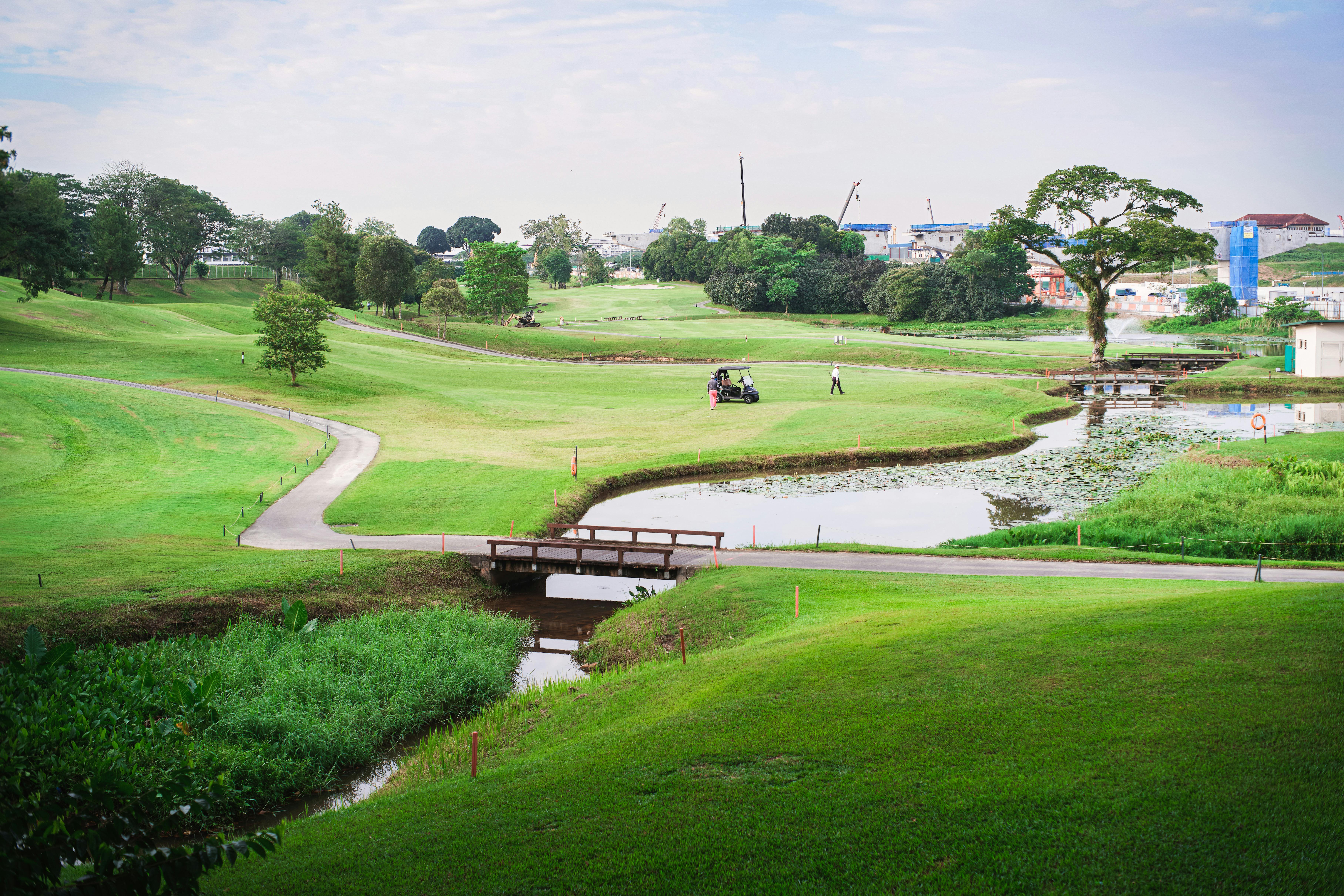 A tranquil view of a lush golf course with a golf cart, bridge, and water feature under a partly cloudy sky.