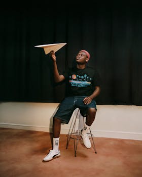 An adult male sitting on a stool indoors, holding a paper airplane, under warm lighting.