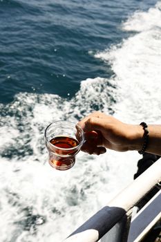 A person holds a glass of tea over a boat railing against the ocean backdrop, enjoying a day at sea.