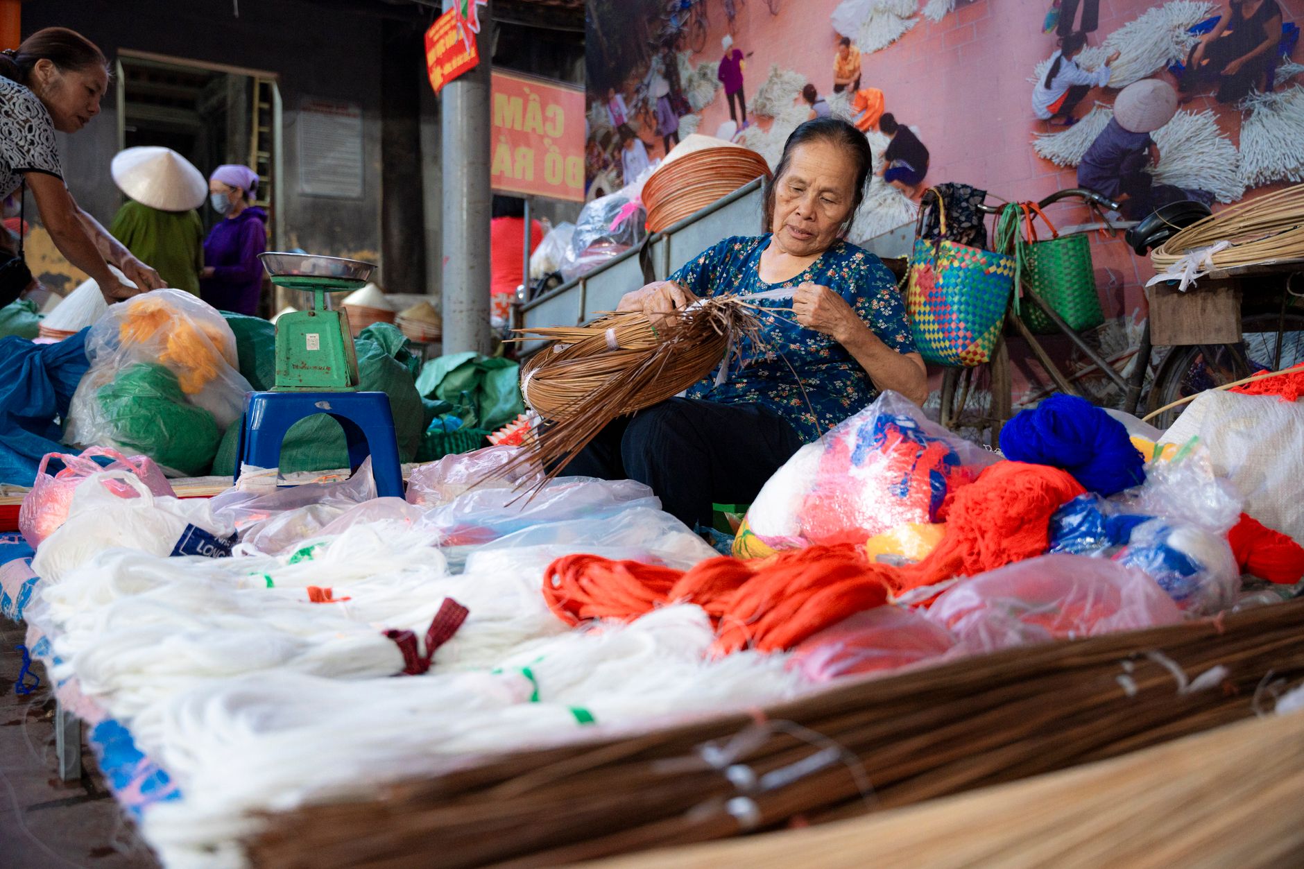 An elderly woman weaving rattan in a vibrant Hà Nội market, surrounded by colorful textiles.