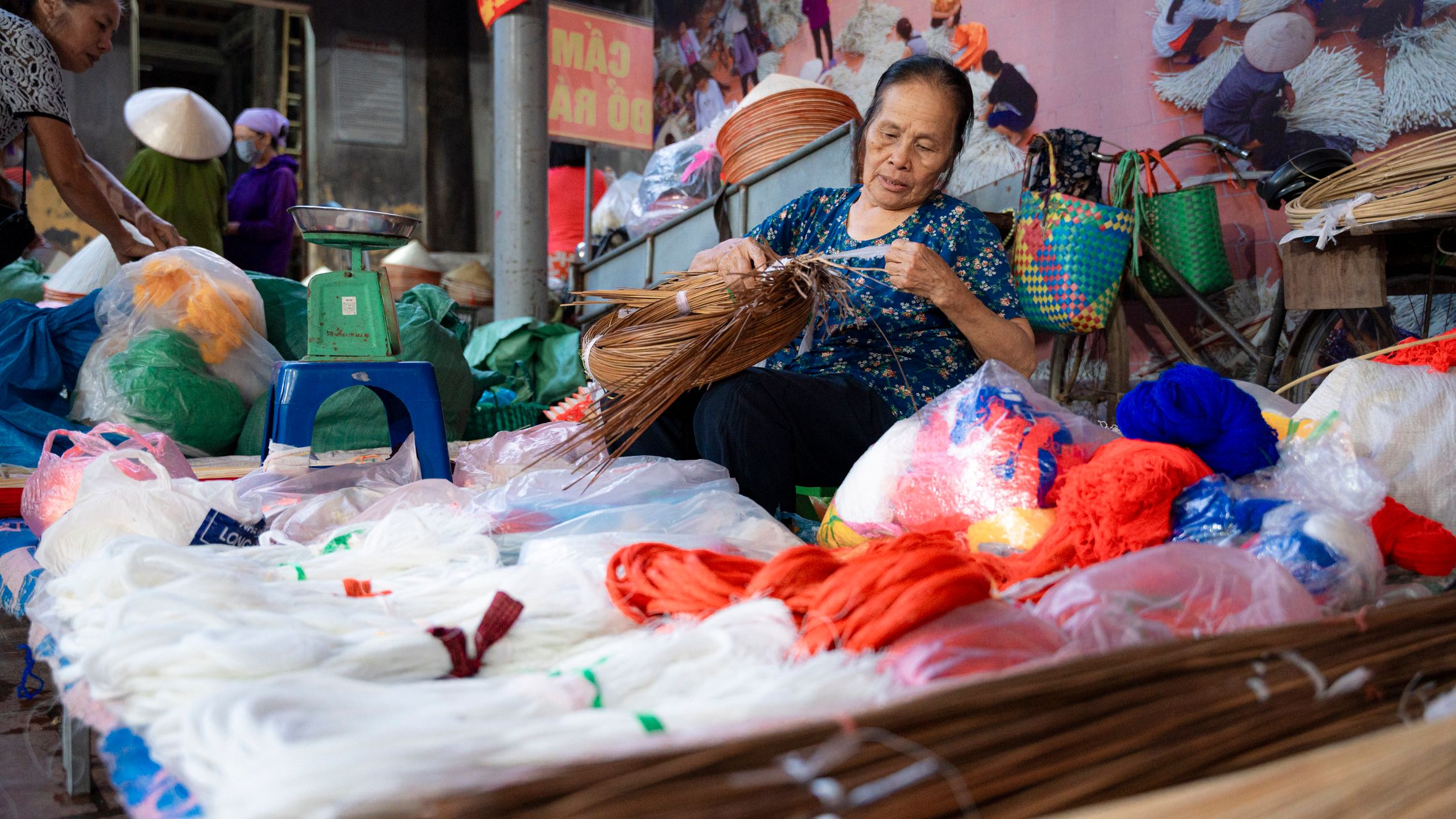 An elderly woman weaving rattan in a vibrant Hà Nội market, surrounded by colorful textiles.