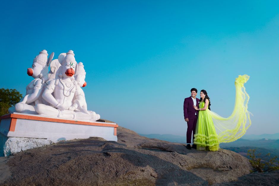 A couple in vibrant attire poses with a striking Hanuman statue at a scenic outdoor location in Hyderabad.