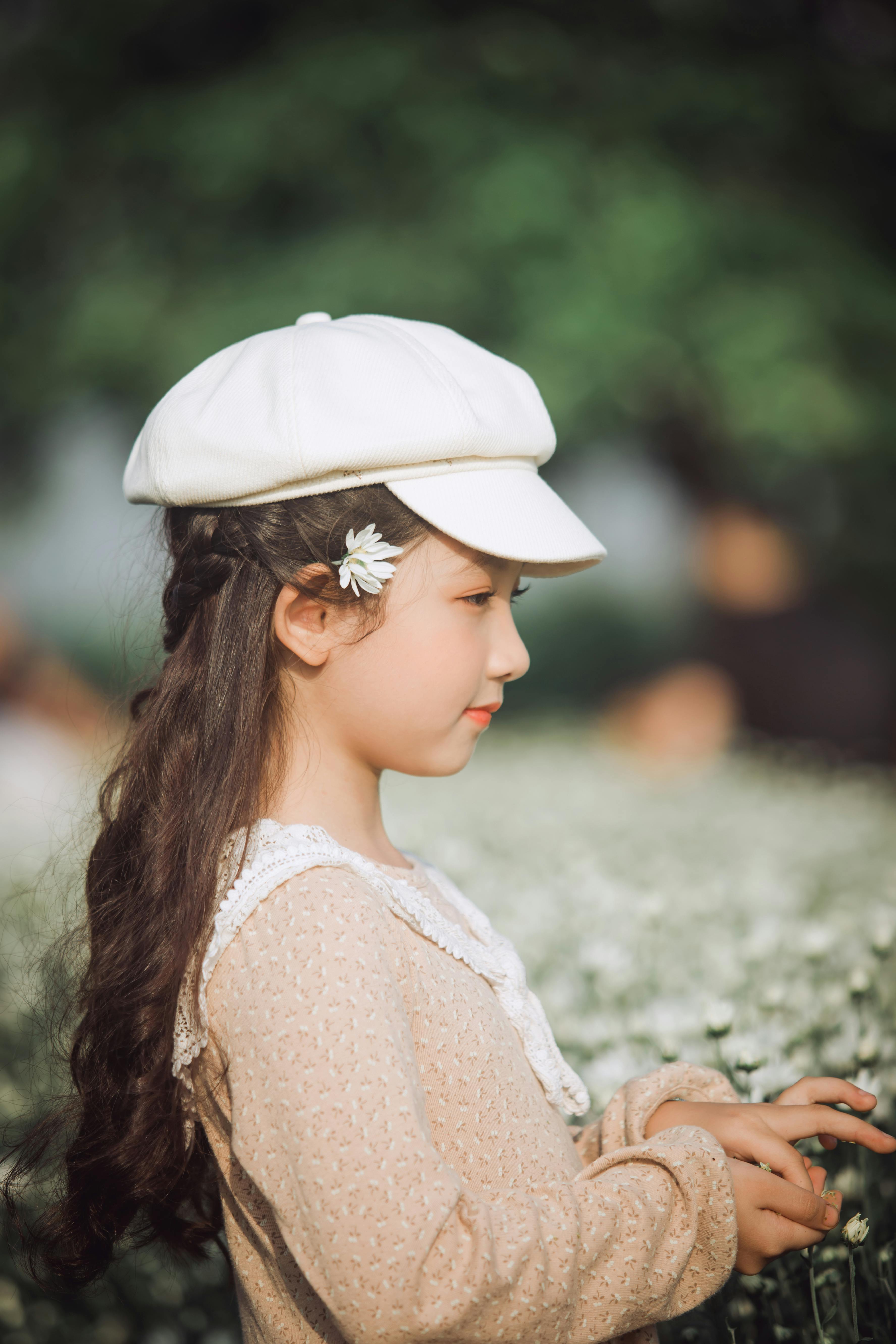 A cute young girl with a white cap and floral dress surrounded by wildflowers outdoors.