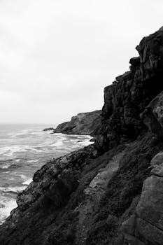 Monochrome view of jagged cliffs by a rugged coastline under an overcast sky.