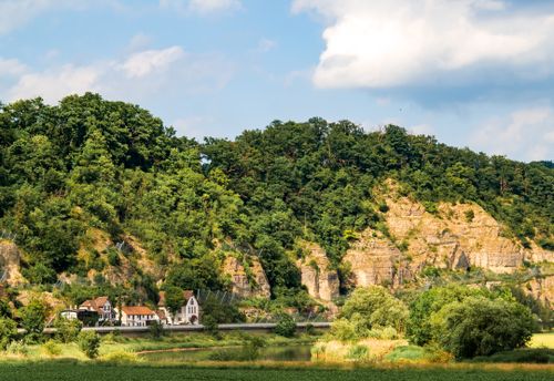 Free Charming riverside village nestled by lush green hills under a blue sky, perfect for travel and nature enthusiasts. Stock Photo